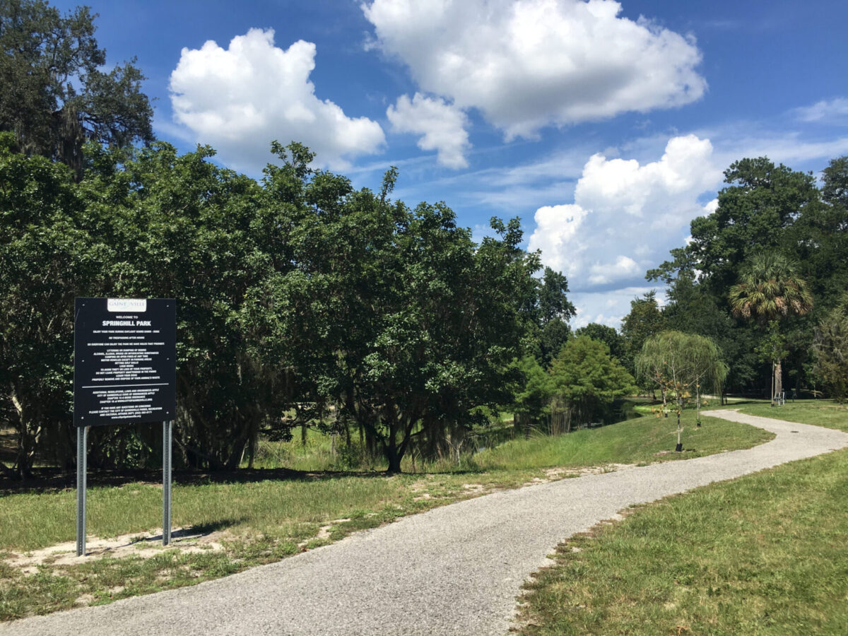 An asphalt pathway weaves between trees under an open sky. A Springhill Park sign stands in the grass-covered-sandy-soil next to the path in the foreground. The grunds next to the path slope down into a waterway.