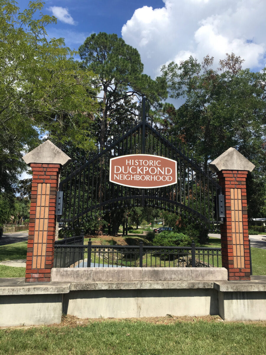 The Sweetwater waterway passes between streets through grassy banks in a neighborhood and under the Historic Duckpond Neighborhood sign.
