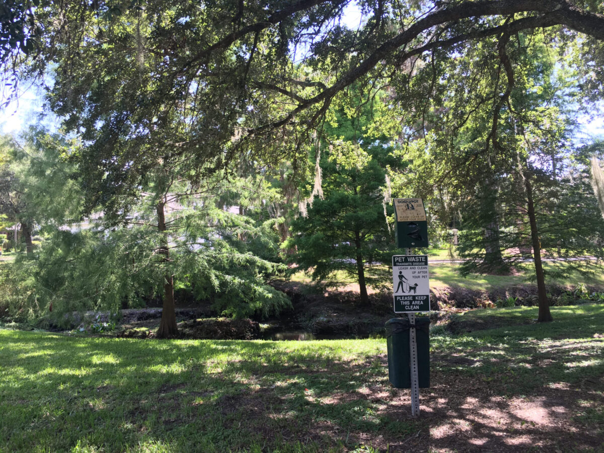 The tree-shaded grassy banks of the Sweetwater waterway pass a pet waste receptacle with bag dispenser.