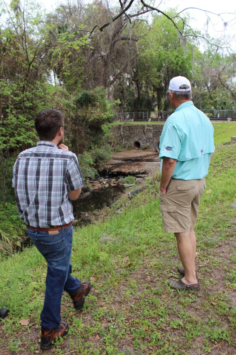 Two men observe a section of Tumblin Creek where the water is coming out from culverts in a stone retaining wall.