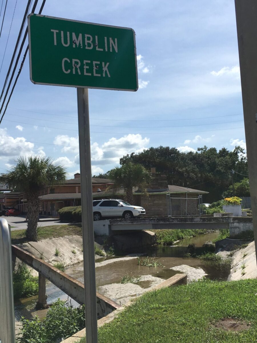 Tumblin Creek Sign in front of a concrete ditch with a small bridge with an SUV crossing it. A hotel can be seen in the background.