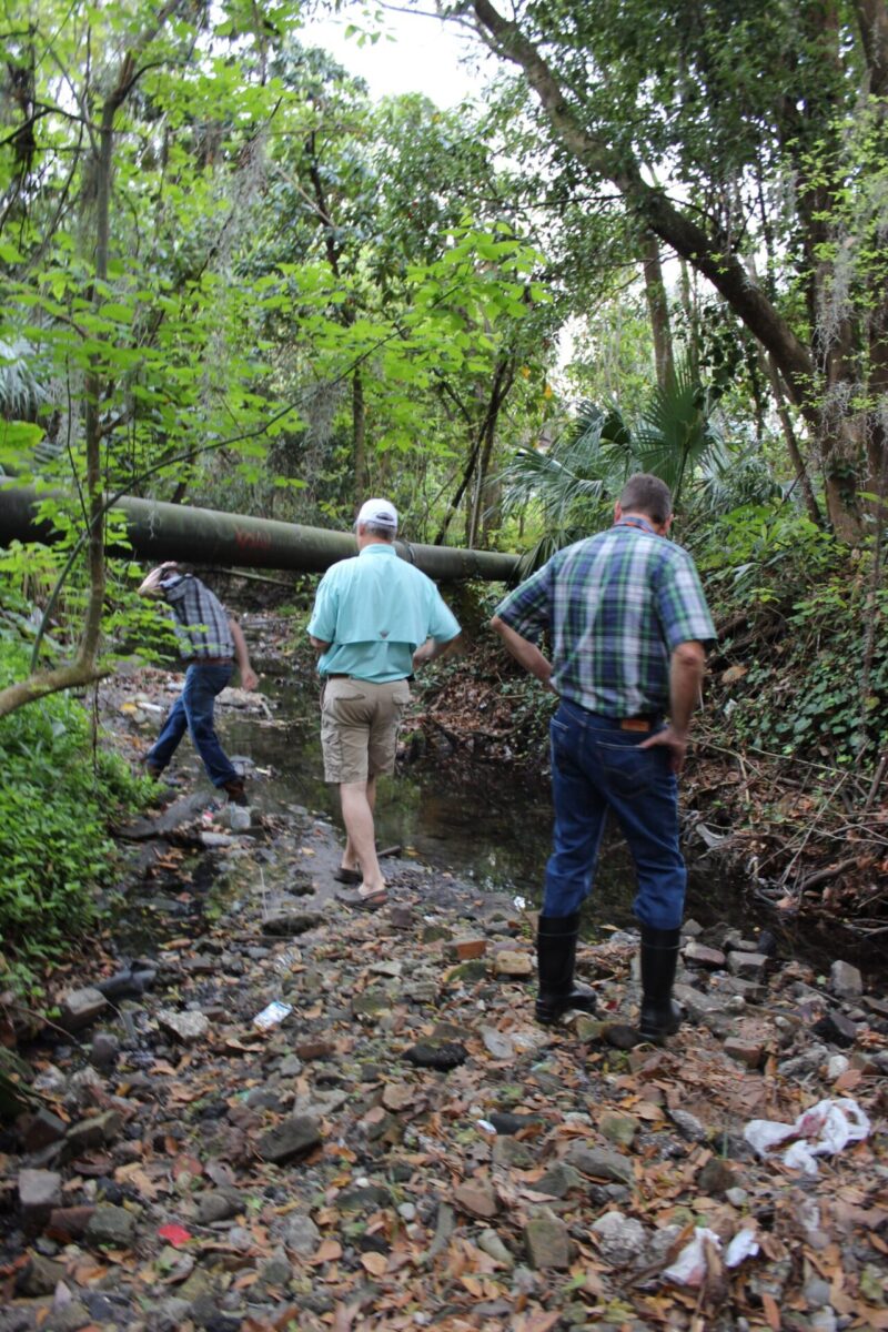 Three men are walking and making observations of Tumblin Creek with a rocky shore. A sewer pipe is suspended across the creek and one of the men is ducking under the pipe as he travels along the creek bank.
