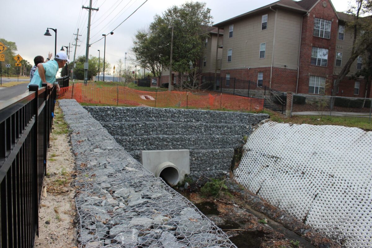 Tumblin Creek passes through a culvert into a rock retaining wall lined with a metal wire mesh. An orange construction fence sits above, with an apartment complex on the right, and a roadway with a sidewalk and short black fence divider to the left of the creek.