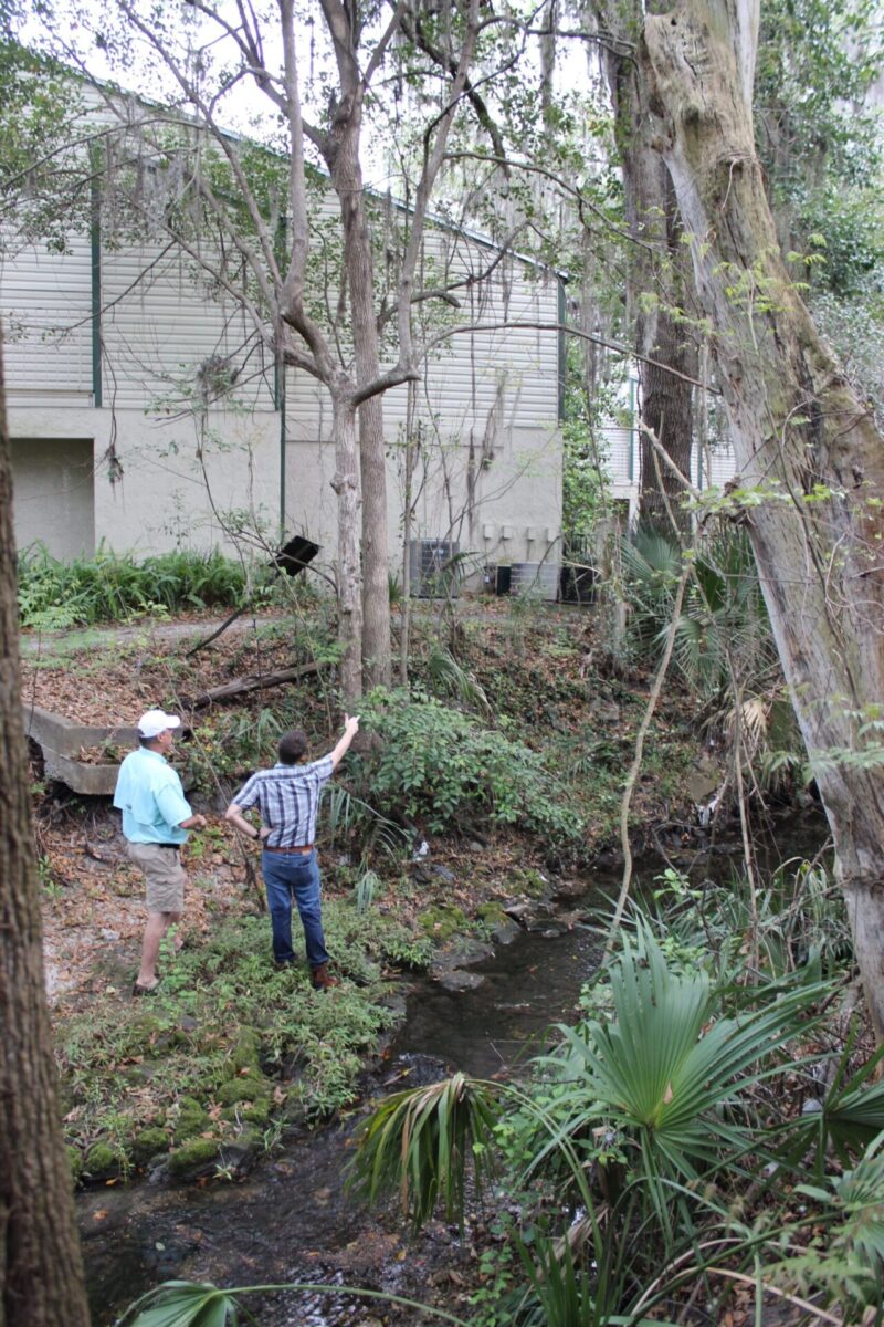 Two men stand on the bank of Tumblin Creek. The men appear to be having a discussion while one of the men points at the trees lining the banks of the creek.